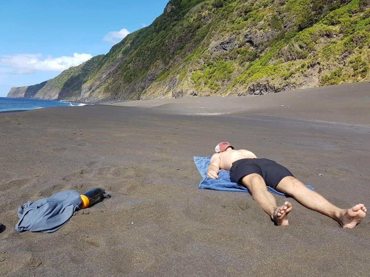 Man sunbathing on black sand beach on Faial in the Azores with cliffs and ocean in the background.