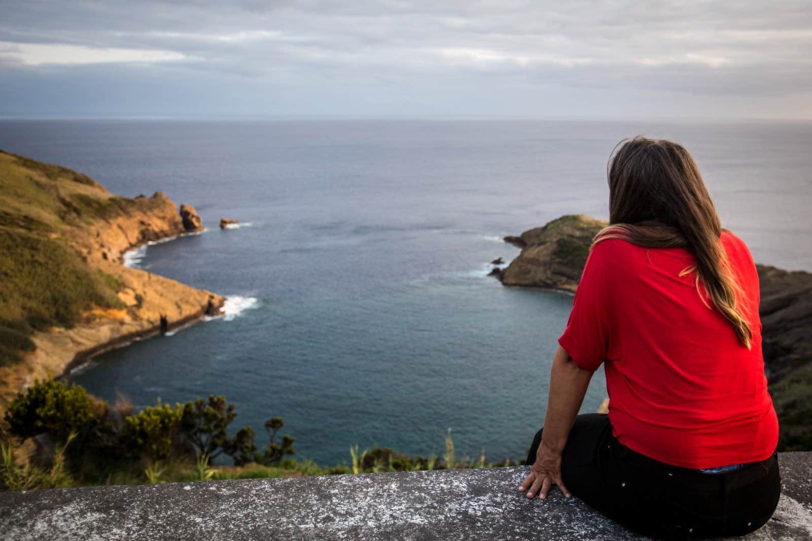 Woman in red shirt sitting on stone wall overlooking a bay on Faial in the Azores