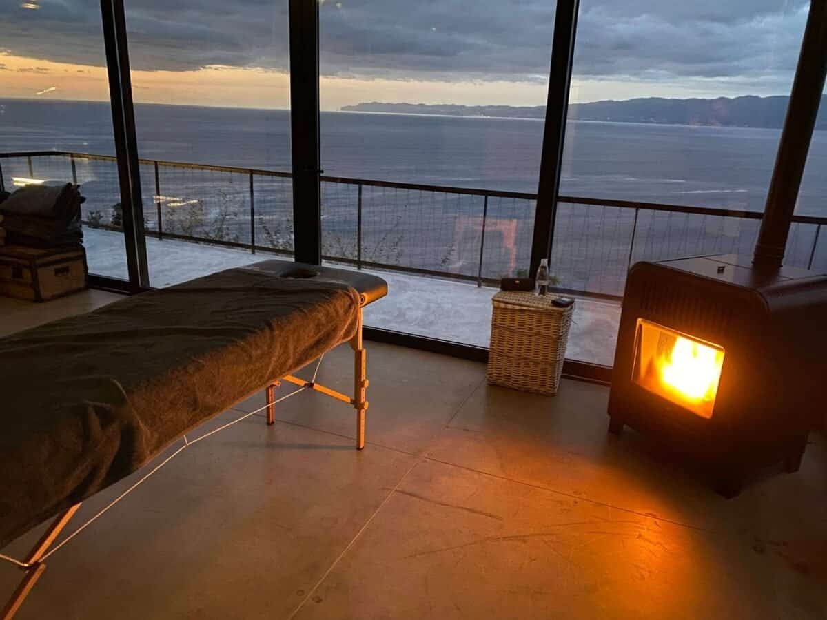 massage table set up next to woodstove and wall-to-floor windows looking out on the ocean
