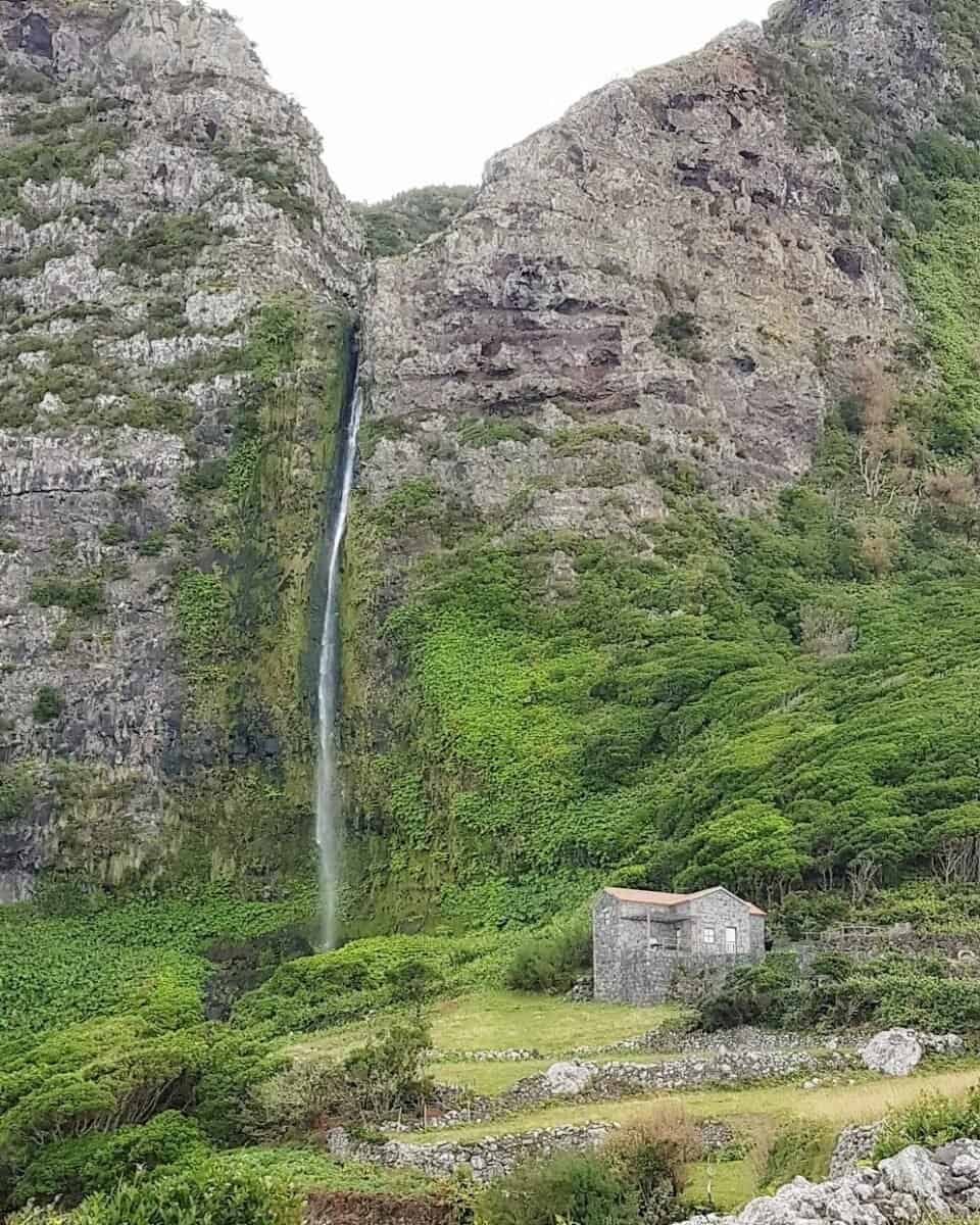 waterfall cascades over tall cliff next to stone house on Flores, Azores